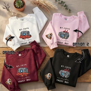 Könnte beinhalten: Vier Sweatshirts in Weiß, Rosa, Burgunderrot und Schwarz, jeweils mit einem grafischen Design. Das Design enthält den Text "BT-7274" über einem stilisierten Bild eines Geräts mit den Worten "TRUST ME!" darunter. Eine kleinere Grafik befindet sich auf dem Ärmel.