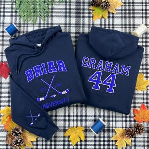 Op de afbeelding: Twee marineblauwe hoodies met witte en blauwe tekst en afbeeldingen. Eén hoodie zegt "Briar University" met gekruiste hockeysticks. De andere zegt "Graham 44". De hoodies hebben trekkoorden en een capuchon.