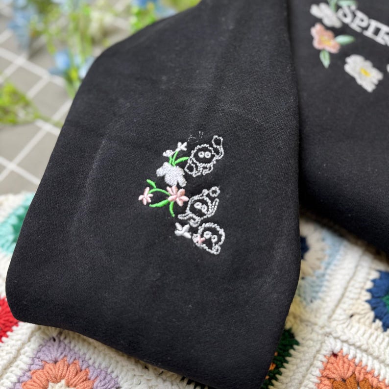 Pode incluir: Artigo de tecido preto com bordados florais e de animais. O bordado apresenta cores brancas, rosa e verdes. A palavra "SPIR" &eacute; vis&iacute;vel em bordado branco no lado direito da imagem. O artigo &eacute; colocado sobre uma superf&iacute;cie colorida e estampada.