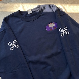 Puede incluir: Una sudadera azul marino de cuello redondo con un gráfico bordado de un personaje en tonos morado y gris en la parte superior derecha del pecho y diseños de eslabones de cadena blancos bordados en ambas mangas. Tiene cuello acanalado.
