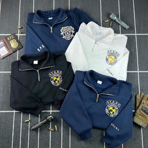 May include: Four quarter-zip sweatshirts in navy, white, and black featuring R.P.D. and S.T.A.R.S. Raccoon Police logos with yellow stars. Includes a brown leather R.P.D. badge wallet, two metal flashlights, several brass bullet casings, and a tactical glove.