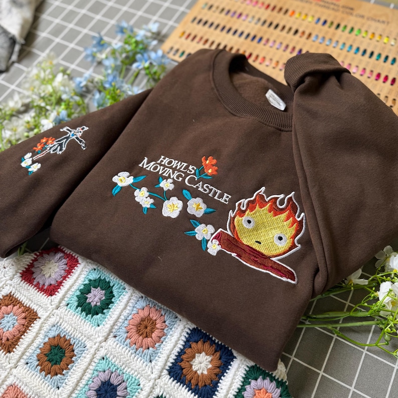 May include: A brown sweatshirt with embroidered floral and character designs. The text "Howl's Moving Castle" is embroidered on the front. The sweatshirt is folded on a colorful, patterned blanket.