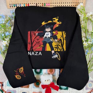 Pode incluir: Uma camisola preta de manga comprida com um bordado central de uma personagem estilo anime rodeada por chamas e caules de bambu, com o texto NAZA por baixo. Um design bordado mais pequeno de duas rodas em chamas aparece na manga.