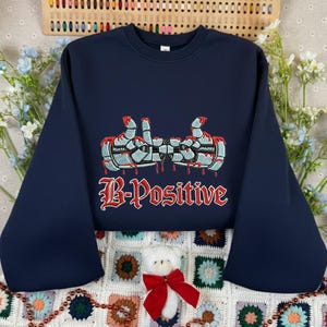 May include: Navy blue sweatshirt with an embroidered design featuring two thumbs up with a dripping blood effect and the text "B-Positive" in red. The sweatshirt has long sleeves and a crew neck.