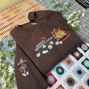 May include: A brown sweatshirt with embroidered designs. The front features text that reads "Howls Moving Castle" with a cartoon character and floral accents. The sleeve has a smaller embroidered design of a character and flowers.