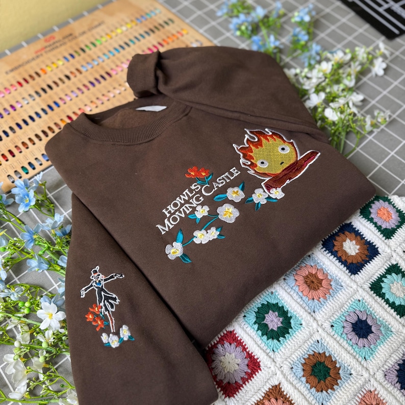 May include: A brown sweatshirt with embroidered designs. The front features text that reads "Howls Moving Castle" with a cartoon character and floral accents. The sleeve has a smaller embroidered design of a character and flowers.