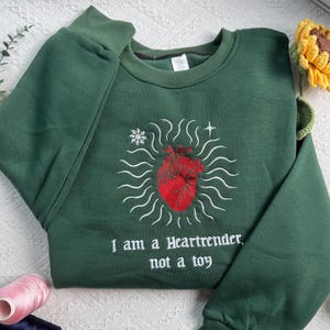 Puede incluir: Una sudadera de cuello redondo verde oscuro con un corazón anatómico rojo bordado en el centro, rodeado de líneas onduladas blancas, un copo de nieve y una estrella. Debajo del corazón, el texto dice: I am a Heartrender, not a toy.