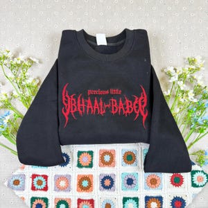 Könnte beinhalten: Schwarzes Sweatshirt mit Rundhalsausschnitt und dem Text "precious little BHAAL BABE" in roter gotischer Schrift. Das Sweatshirt wird auf einer bunten, gehäkelten Decke mit floralen Akzenten präsentiert.