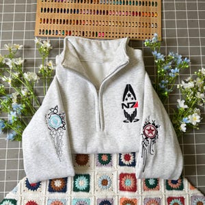 Puede incluir: Sudadera gris claro con cremallera y diseños bordados en las mangas y el pecho. El pecho presenta un emblema negro y rojo con las letras "NZ". Las mangas tienen diseños intrincados y coloridos.