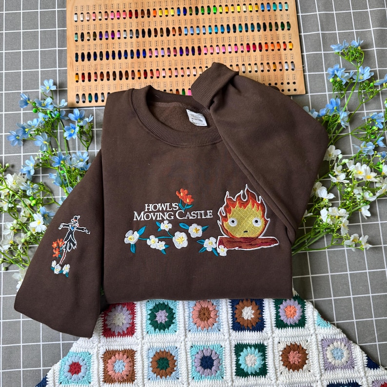 May include: A brown sweatshirt with embroidered designs from "Howl's Moving Castle." The design includes the text "Howl's Moving Castle," a fire demon, flowers, and a character on the sleeve. The sweatshirt is displayed on a checkered surface.