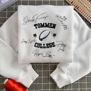 May include: A white long-sleeve sweatshirt featuring black embroidered text that reads TOMMEN COLLEGE. The design includes a central rugby ball graphic, two small green shamrocks, and several handwritten-style signatures in black thread.