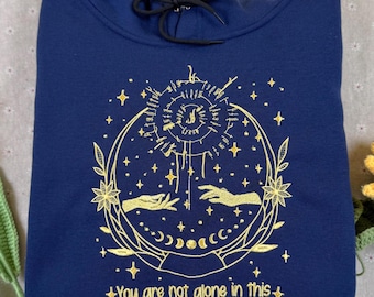 May include: A navy blue hooded sweatshirt featuring a gold embroidered design of two hands reaching toward a central sun-like symbol, surrounded by stars, moon phases, and floral elements, with the text "You are not alone in this".