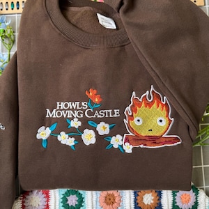 May include: A brown sweatshirt with embroidered artwork from "Howl's Moving Castle." The design includes the text "HOWL'S MOVING CASTLE" with floral and fire demon imagery. The sweatshirt is made of a soft material.