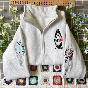 Puede incluir: Una sudadera gris jaspeado con cremallera de un cuarto y el logotipo N7 bordado en el pecho izquierdo. Las mangas presentan intrincados bordados de estilo circuito en negro, azul y rojo con un emblema de estrella en la manga derecha.