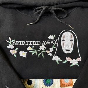 Pode incluir: Moletom preto com um design bordado do filme "A Viagem de Chihiro". O design inclui o texto "Spirited Away" com detalhes florais e uma ilustra&ccedil;&atilde;o de personagem. O moletom tem um cord&atilde;o e ponteiras de metal.