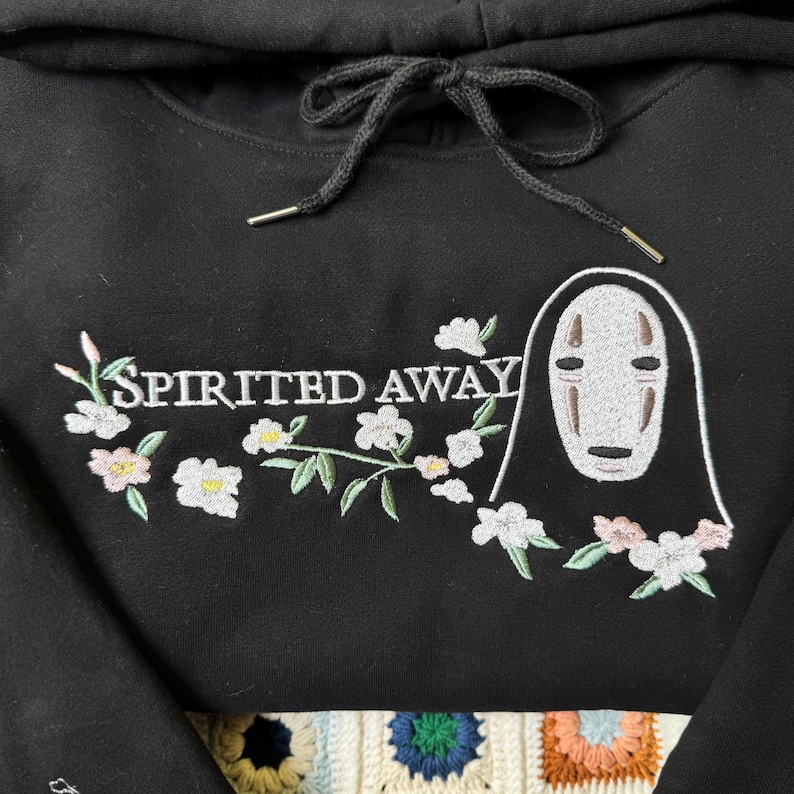 Pode incluir: Moletom preto com um design bordado do filme "A Viagem de Chihiro". O design inclui o texto "Spirited Away" com detalhes florais e uma ilustra&ccedil;&atilde;o de personagem. O moletom tem um cord&atilde;o e ponteiras de metal.