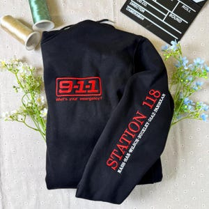 Puede incluir: Sudadera negra con texto bordado en rojo. La parte delantera presenta "9-1-1" dentro de un rectángulo rojo, con "What's your emergency?" debajo. La manga muestra "STATION 118" y nombres. Sobre una superficie blanca con flores y costura.