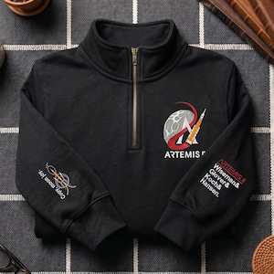 Pode incluir: Camisola preta com fecho de correr de um quarto de zip dourado. A camisola apresenta um design bordado de um foguete e da lua com a palavra "ARTEMIS" e o texto "ARTEMIS II Wiseman & Glover & Koch & Hansen." A manga esquerda tem o texto "Copy, moon joy."
