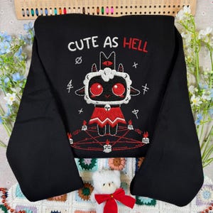 May include: Black sweatshirt with the embroidered phrase "CUTE AS HELL" and a cartoon lamb design. The lamb has red eyes and a red outfit, surrounded by candles and symbols. A unique and eye-catching apparel item.