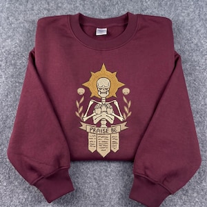 Peut inclure: Un sweat-shirt à col rond bordeaux avec une broderie centrale représentant un squelette doté d'une auréole dorée, entouré de deux petits crânes et de branches feuillues. Sous le squelette, une bannière porte l'inscription "PRAISE BE" et trois parchemins.