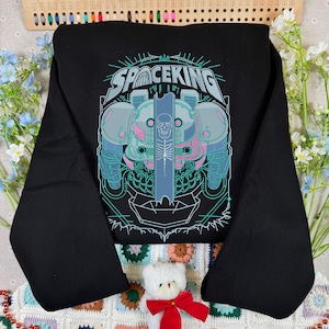 May include: Black sweatshirt with a graphic design featuring the text "SPACEKING" above a detailed illustration of a futuristic robot-like figure with a skull and skeleton design, in shades of teal, pink, and blue.