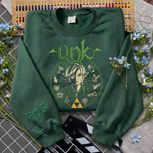 May include: A forest green crewneck sweatshirt featuring an embroidered graphic of a character with pointed ears surrounded by various small icons and the text Link. The right sleeve displays an embroidered winged triangle emblem in green thread.