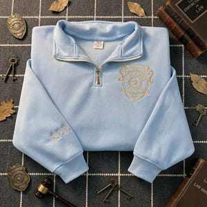 May include: A light blue quarter-zip sweatshirt featuring a gold embroidered badge emblem on the left chest with the text Springfield Police and a star, and the letters B.P.U. embroidered in gold on the right sleeve cuff with a silver zipper.