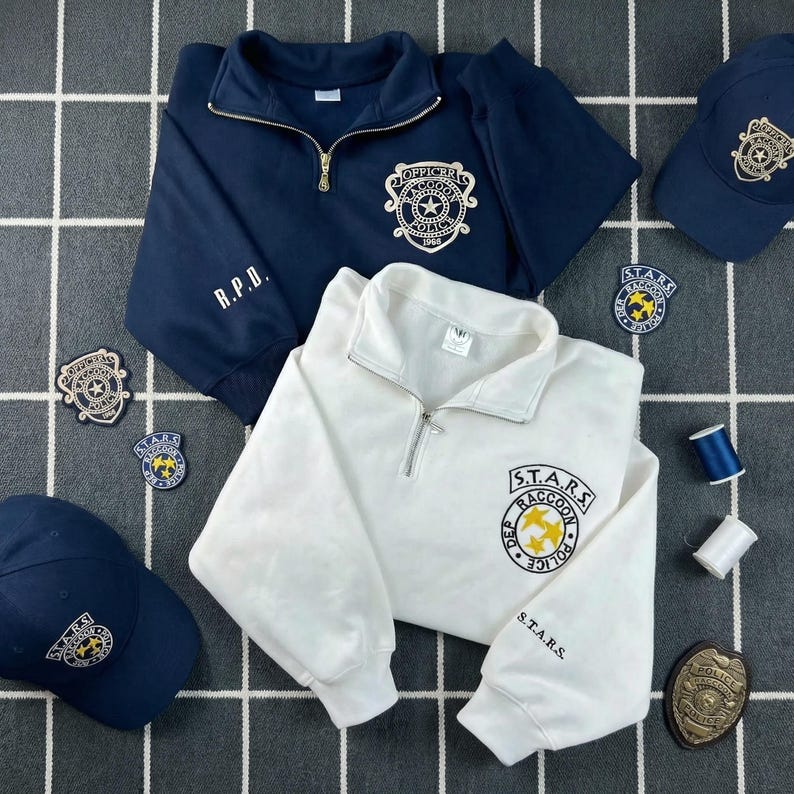 May include: A navy blue quarter-zip sweatshirt with R.P.D. and an Officer Raccoon Police emblem, a white quarter-zip sweatshirt with S.T.A.R.S. Raccoon Police emblem, two baseball caps with emblems, three embroidered patches, and a gold-toned badge.