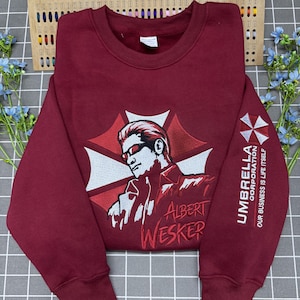 Pode incluir: Uma camisola de gola redonda cor de vinho com um gráfico bordado de Albert Wesker em frente ao logótipo vermelho e branco da Umbrella Corporation. A manga apresenta o logótipo da Umbrella Corporation e o texto, "OUR BUSINESS IS LIFE ITSELF."