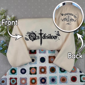 Può includere: Felpa color crema con testo e grafica ricamati in nero. La parte anteriore presenta la parola "Quicksilver" con una spada e una farfalla. Il retro ha "Kingfisher" con un disegno di montagna e farfalla. La felpa è su una colorata coperta all'uncinetto.