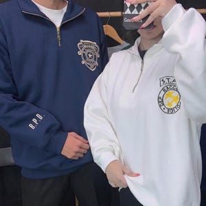 May include: A navy blue quarter-zip sweatshirt with a gold Raccoon Police Officer patch and R.P.D. text on the sleeve, alongside a white quarter-zip sweatshirt featuring a S.T.A.R.S. Raccoon Police patch with a yellow star emblem on the chest.