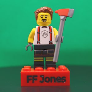 May include: A Lego minifigure with brown hair, a mustache, and a white shirt featuring red suspenders and a flame emblem. The figure wears black pants with yellow reflective stripes, holds a gray and red axe, and stands on a red brick labeled FF Jones.