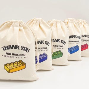 Può includere: Cinque piccole borse di cotone bianco con coulisse, con la scritta "Grazie per aver costruito ricordi con me!" stampata su di esse. Ogni borsa presenta un mattoncino Lego di colore diverso.