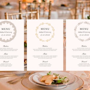 May include: Three white wedding menu cards with gold and silver floral wreaths. The cards list the menu for Aisha & Farooq's wedding on 15 October 2022. The menu includes starters, main course, and dessert.