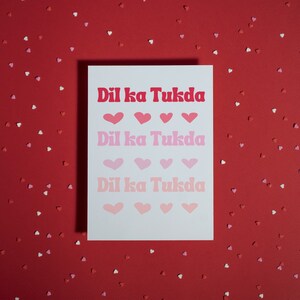 May include: A white card with the text "Dil ka Tukda" repeated three times in pink, red, and light pink. The text is in a retro font style. The card is surrounded by small pink, red, and white hearts on a red background.