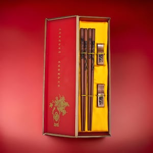 Op de afbeelding: Een set houten eetstokjes in een rode en gouden doos. De eetstokjes zijn versierd met zwarte en gouden ontwerpen. De doos heeft Chinese karakters erop.