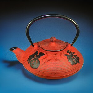 May include: Red cast iron teapot with a black handle and lid. The teapot has black Japanese characters on the side.