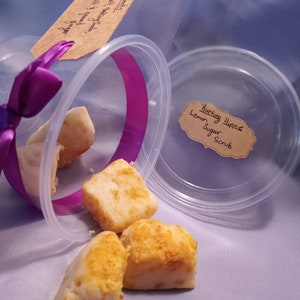 May include: Four white sugar scrub cubes with a yellow citrus zest topping, in a clear plastic container with a purple ribbon. The label reads "Riveting Hippie Lemon Sugar Scrub".