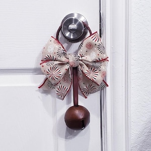 Puede incluir: Una campana de metal marrón con un lazo de cinta rojo y blanco atado alrededor del mango. El lazo tiene un patrón de fuegos artificiales rojos y blancos sobre un fondo beige.