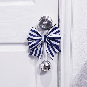 Puede incluir: Una puerta blanca con un lazo azul y blanco a rayas atado alrededor del pomo de la puerta. Una campanilla plateada cuelga del lazo.