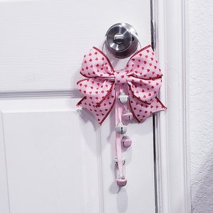 May include: A pink bow with red hearts printed on it is tied to a door handle. A string of silver bells hangs from the bow.
