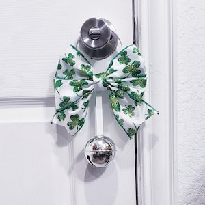 May include: A white door with a silver doorknob is adorned with a St. Patrick's Day decoration. A large bow made of white fabric with green shamrocks and glitter is attached to a silver bell. The bell hangs from a white ribbon.