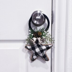 Puede incluir: Un adorno en forma de estrella a cuadros blanco y negro con campanas plateadas y vegetación colgando de un pomo de puerta blanco.
