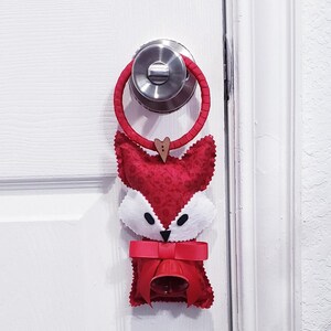 Può includere: Un appendiporta a forma di volpe rossa con un viso bianco e una campana appesa a un fiocco rosso. L'appendiporta è attaccato a una porta bianca con una maniglia argentata.