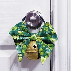 May include: A gold bell with a star-shaped cut-out hangs from a door handle. It's decorated with a green bow patterned with shamrocks, ideal for St. Patrick's Day celebrations.