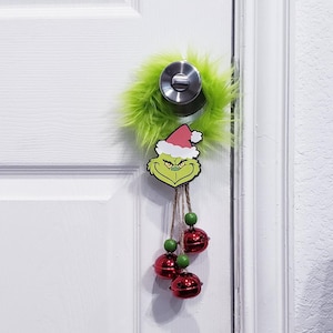 以下が含まれることがあります： 白いドアに、銀色のドアノブが付いている。緑色のふわふわしたリースがドアにかけられており、リースには緑色の毛皮と赤いサンタ帽子のグリンチの顔が描かれている。リースには赤い鈴がぶら下がっている。