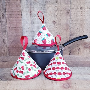 Puede incluir: Tres agarraderas de olla acolchadas en forma de cono con ribete rojo. Las agarraderas están decoradas con un fondo blanco y cerezas o fresas rojas. Las agarraderas están sobre una superficie de madera.