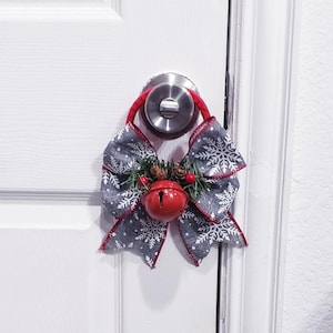 Può includere: Un appendino per porta natalizio rosso e bianco con una campana e un fiocco. Il fiocco è fatto di tessuto grigio con fiocchi di neve bianchi stampati su di esso. L'appendino è attaccato a una maniglia della porta.