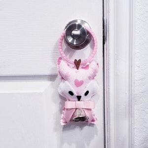 May include: A pink fox-shaped doorstop with a bell and a pink bow. The doorstop is hanging on a white door.