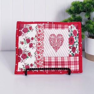 Puede incluir: Un posavasos acolchado rojo y blanco con un dise&ntilde;o de coraz&oacute;n. El posavasos est&aacute; hecho de tela con un patr&oacute;n de patchwork de corazones, flores y cuadros. Se muestra sobre una superficie blanca con una peque&ntilde;a planta en maceta en el fondo.